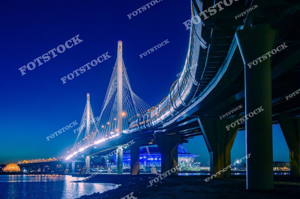 На фотографии изображен Большой Обуховский мост, часть Западного скоростного диаметра (ЗСД), в ночном освещении. Элегантные вантовые конструкции моста подчёркнуты яркой подсветкой, создавая впечатляющую картину на фоне тёмного неба. На заднем плане виднеется стадион, добавляющий современный городской акцент. Это инженерное сооружение является важной транспортной артерией города, соединяющей берега Финского залива.